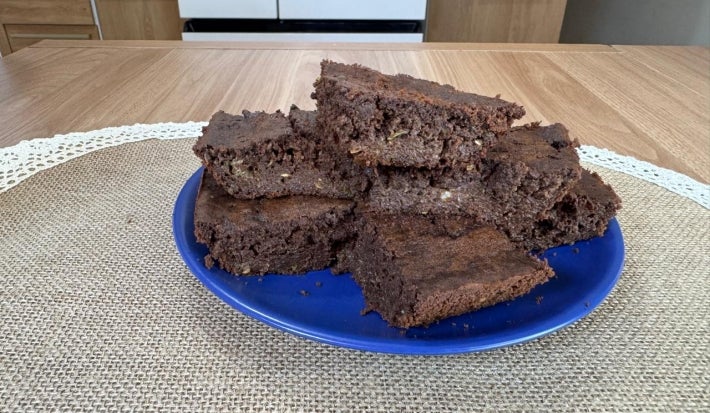Fotografia de um prato raso azul sobre um paninho de tom bege apoiado em uma mesa de madeira. Sobre o prato tem alguns pedaços empilhados do brownie de abobrinha.