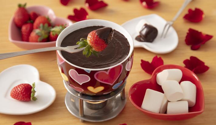 Fotografia em tons de vermelho em uma bancada de madeira de cor marrom. Ao centro, um richaud contendo o fondue. Ao redor, há 2 pratos brancos contendo morangos, 1 prato vermelho contendo marshmallow e 1 pote rosa contendo morangos.