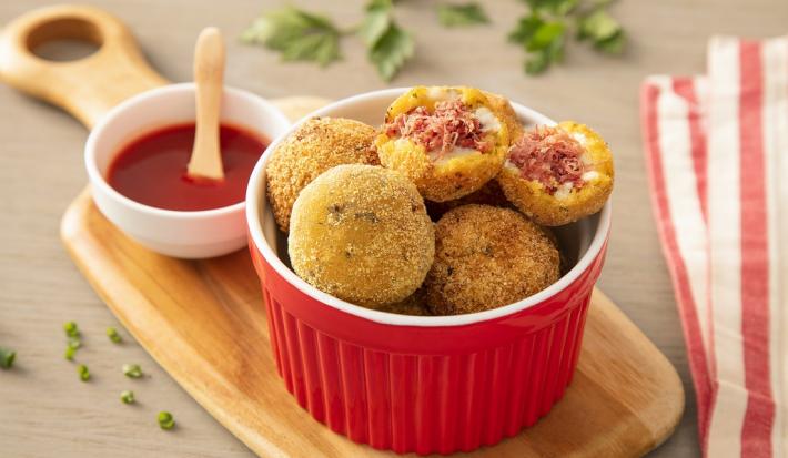 Fotografia em tons de amarelo, em uma bancada de madeira de cor marrom. AO centro, uma tábua de madeira com um recipiente vermelho contendo os bolinho de carne. Ao fundo, um pano listrado vermelho, um pote de katchup e folhas de salsinha espalhadas.