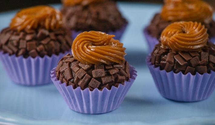 Fotografia de cinco unidades de forminhas azuis com brigadeiro de pão de mel, com doce de leite por cima de cada unidade. Os brigadeiros estão sobre um apoio decorativo de cor azul.