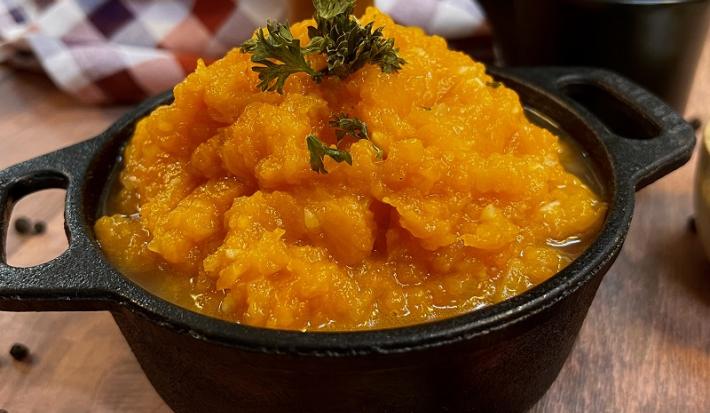 Foto em tons de laranja da receita de quibebe servida em uma panela de pedra preta com folhas salsinha para decorar. Ao fundo há um paninho quadriculado branco e vermelho além de uma xícara preta, tudo isso sobre uma mesa de madeira