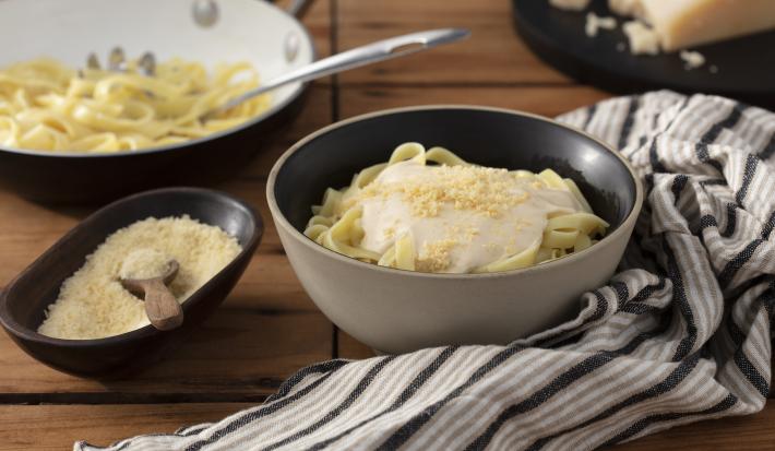 Fotografia em tons de marrom em uma bancada de madeira escura, um pano branco com listras marrom, um recipiente redondo fundo bege com o fettuccine com molho alfredo e queijo ralado por cima dentro dele. Ao lado, um potinho com queijo ralado.