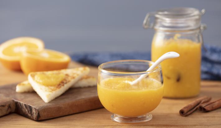 Fotografia em tons de laranja em uma bancada de madeira, uma tábua de madeira, um paninho azul, um pote hermético de vidro com geleia de laranja dentro. Ao lado, um potinho de vidro com mais geleia. Ao lado, fatias de pão e laranjas cortadas ao meio.