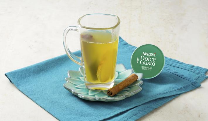 Fotografia em tons de cinza e azul de uma bancada vista de frente. Contém um pano azul, um pratinho em formato de rosa com uma xicara transparente que contém a bebida. Ao lado uma capsula de Dolce Gusto.