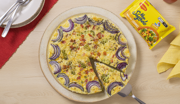 Fotografia mostra a Torta de Legumes dourada e fofinha, com pedaços de legumes visíveis na superfície, servida em um prato.