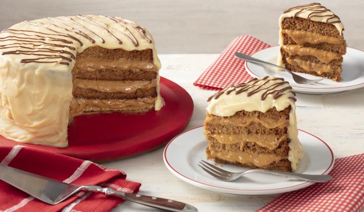 Fotografia em tons de vermelho em uma bancada cinza com paninhos vermelhos listrados e de bolinhas brancas. Ao centro, um prato branco com uma fatia do bolo de pão de mel com chocolate branco e à esquerda, um suporte grande vermelho com o bolo inteiro.