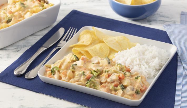 Fotografia em tons de azul em uma bancada de mármore branco com um jogo americano azul escuro, um prato quadrado branco grande com o strogonoff de legumes, arroz branco e batatas chips dentro dele. Ao fundo, um recipiente com mais strogonoff de legumes.