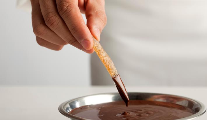 Fotografia de fruta seca afundando no chocolate amargo, que está em um recipiente de alumínio fundo. O qual está sobre uma mesa branca.
