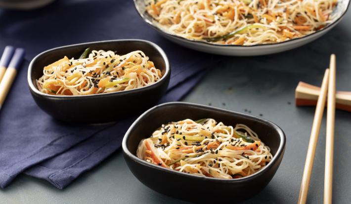 Fotografia em tons de azul escuro em uma bancada de madeira cinza escura com três recipientes com a salada de macarrão tipo bifum, comida japonesa com um hashi ao lado.