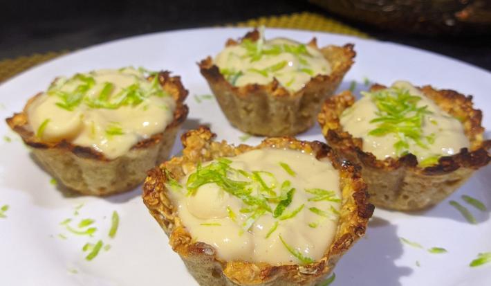Foto aproximada de quatro mini tortinhas de limão, numa base em tom marrom claro e com um recheio cremoso, decoradas com raspas de limão em um prato branco