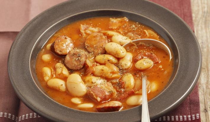 Fotografia de um prato fundo na cor preta com uma sopa de feijão branco e linguiça. Ao lado, três fatias de pão e do outro lado, um tempero em pó vermelho dentro de um pote pequeno branco de cerâmica, que está em cima de um pano em tons de vinho.