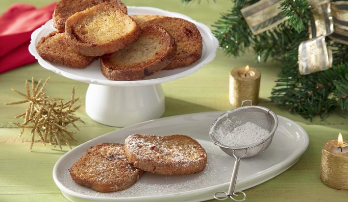 Foto da receita de rabanada sem glúten servida sobre uma base de porcelana branca com duas fatias separadas polvilhadas com açúcar de confeiteiro. Ao fundo há um paninho vermelho e decoração natalina