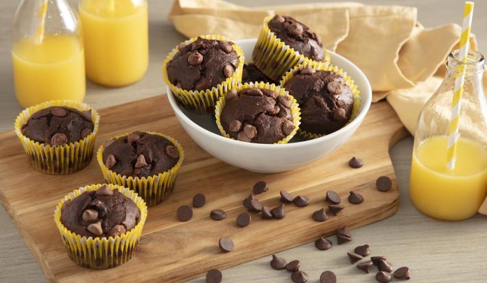 Fotografia em tons de amarelo em uma bancada de madeira com um pano bege, uma tábua de madeira ao centro, com os mini bolinhos de chocolate com gotas de chocolate em forminhas amarelas em cima da tábua e um pote branco com mais bolinhos.