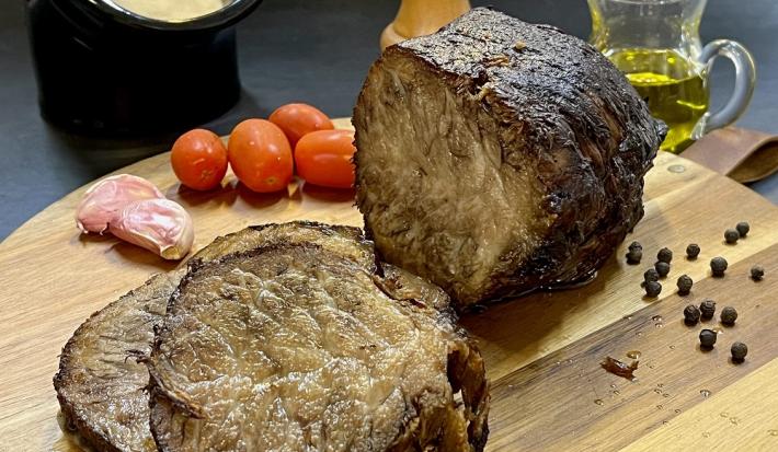 Foto em tons de marrom da receita de cupim na pressão servida em uma tábua de madeira com a peça de cupim cortada em algumas fatias e com tomates cerejas e pimentas. Ao fundo há uma jarra de vidro com azeite além de um recipiente preto com molho branco
