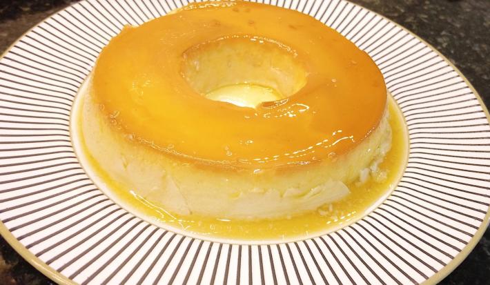Foto aproximada de um pudim de leite condensado em tom amarelo, num prato redondo branco com decoração listrada. Na imagem há um selo em tom azul claro com o texto Receita do Consumidor.