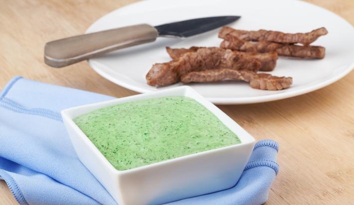 Fotografia em tons de azul e verde em uma bancada de madeira clara, um pano azul, um potinho quadrado branco com o patê cremoso de chimichurri dentro. Ao fundo, um prato branco redondo com tirinhas de carne para acompanhar o patê.