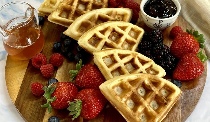 Foto em tons de bege e marrom da receita de waffle delícia servida em seis porções sobre uma tábua de madeira, com frutas vermelhas espalhadas pela tábua, além de um recipiente com geleia de frutas e vidro de mel