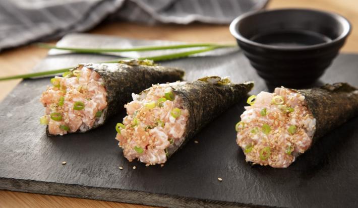 Foto da receita de Temaki de Salmão. Observa-se 3 temakis em cima de uma tábua de preta e, atrás, um potinho com molho shoyu e um ramo de cebolinha.