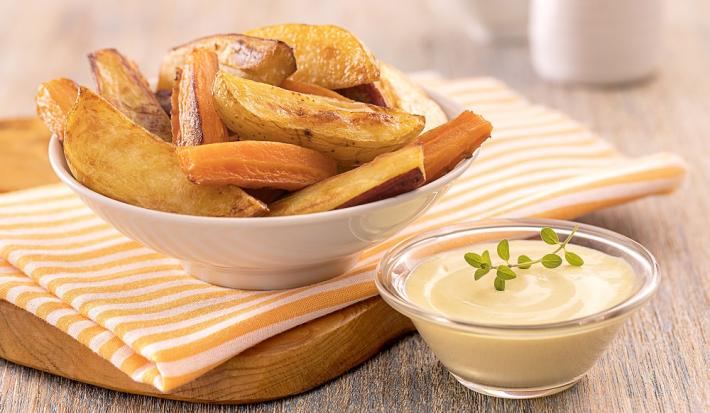 Foto em tons de amarelo da receita de legumes rústicos com molho rosé servida em um bowl de porcelana branco sobre um pano listrado laranja em cima de uma tábua de madeira. À frente há um recipiente de vidro com o molho rosé