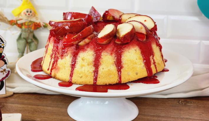 Foto da receita de bolo de maçã do amor, servido em um prato alto branco, com calda vermelha de maçã do amor escorrendo pelas laterais, com pedaços de maçã decorando o topo e itens de festa junina sobre a bancada ao lado do bolo