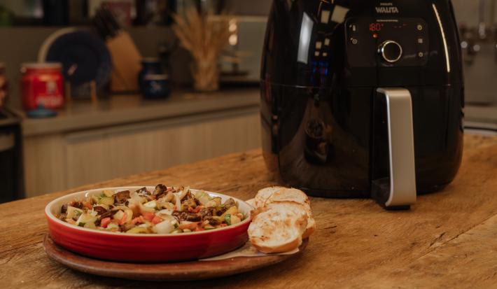Fotografia em tons de marrom de uma bancada de madeira com uma tabua de madeira, sobre ela um recipiente redondo vermelho com a caponata de legumes. Ao fundo uma airfryer.