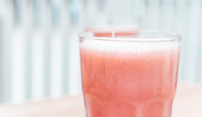 Fotografia em tons claros de uma bebida com um pouco de espuma de melancia com leite MOÇA e neston, que está em um copo de vidro.