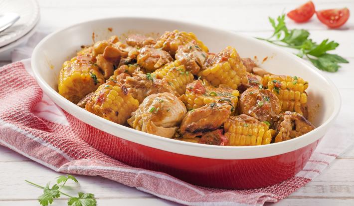 Fotografia em tons de amarelo, vermelho e branco de uma bancada de madeira branca com paninho vermelho, sobre ele um recipiente oval vermelho com bordas brancas com pedaços de frango e milho. Ao fundo duas fatias de tomate e um prato redondo branco.