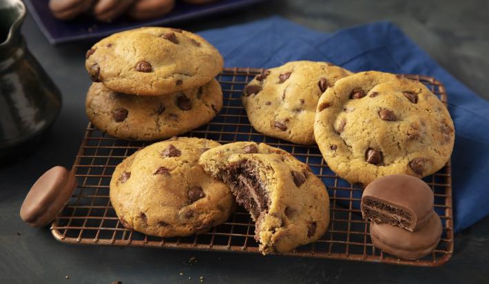 Fotografia em tons de preto e azul de uma bancada preta com um grade dourada, sobre ela cookies. Ao fundo um paninho azul e biscoitos negresco cobertos de chocolate espalhados.