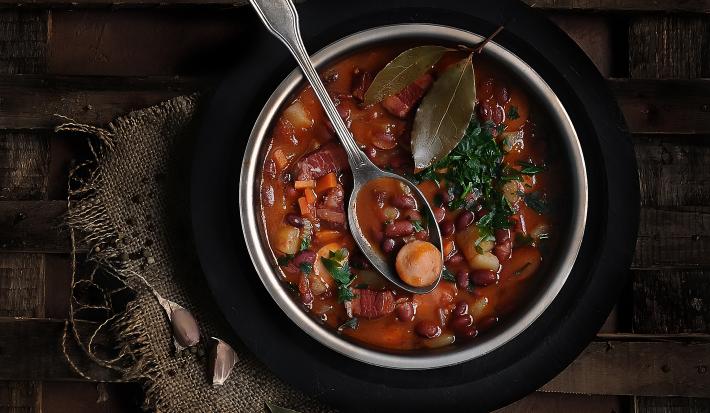 Fotografia vista de cima de uma sopa com linguiça, feijão, cenoura, costela e duas folhas de loura por cima. Apoiada na sopa, uma colher de sopa, que está dentro de um recipiente fundo de alumínio, sobre um prato preto, que está sobre uma mesa de madeira.