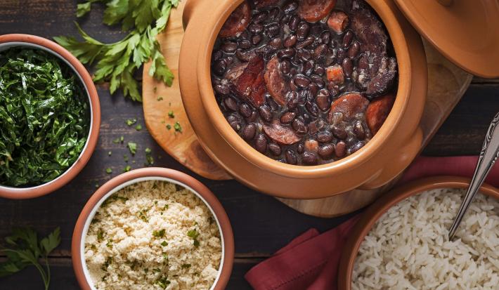 Fotografia em tons de preto e marrom de uma bancada de madeira escura, um pote com couve refogada, um pote com farofa, um pote com arroz branco e um pote com uma feijoada com feijão preto e várias carnes.