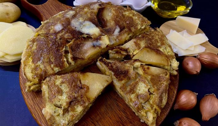Foto em tons de amarelo da receita de tortilla de batata servida em algumas porções sobre uma tábua circular de madeira sobre uma mesa azul. Ao lado há algumas cebolas, fatias de queijo e batatas cortadas em rodelas e inteiras, além de ovos e azeite.