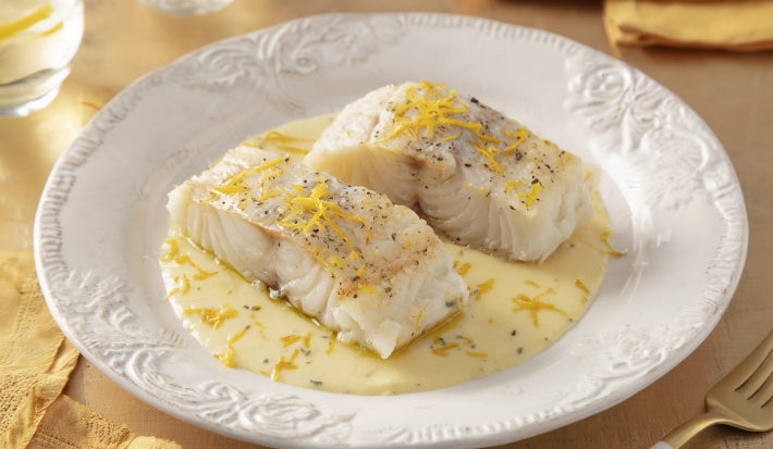 Filés de bacalhau servidos com molho cremoso de ervas e raspas de limão, apresentados em prato branco elegante.
