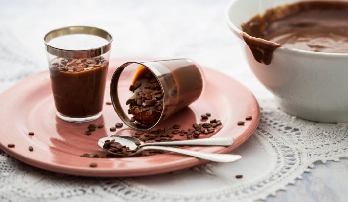 Foto em tons branco e rosa, ao centro prato rosa com dois copinhos contendo brigadeiro e duas colheres de sobremesa, ao lado pote com brigadeiro, sobre tolha branca em bancada de madeira.
