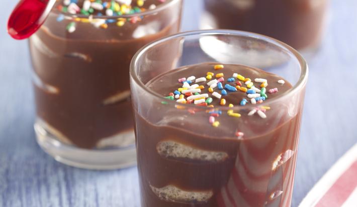 Fotografia em tons de chocolate e azul, com três copinhos com pavê decorados com confeitos coloridos, colheres vermelha e amarela, ao lado guardanapo com listras branca e vinho e colher azul, tudo sobre bancada de madeira azul.