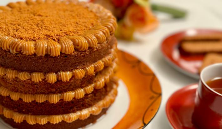 Fotografia em tons de caramelo com um prato de cor marrom ao centro. Em cima do prato existe um bolo com várias camadas de doce de leite e coberto com biscoito triturado