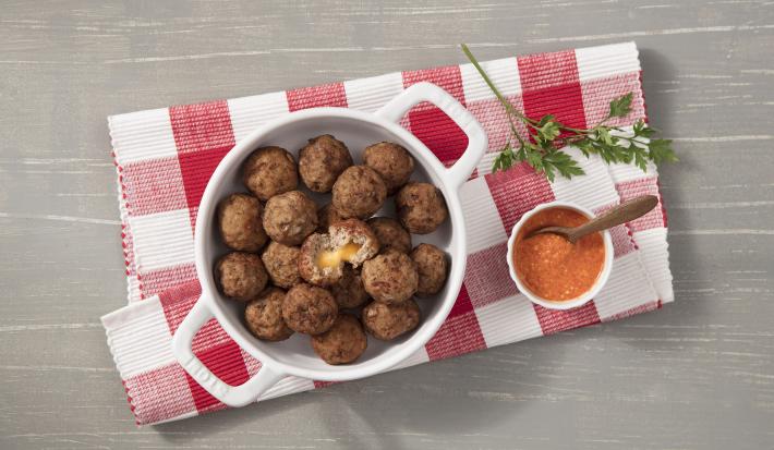 Fotografia em tons de cinza, branco e vermelho de uma bancada cinza vista de cima, ao centro um pano xadrez nas cores vermelho e branco e por cima uma panela branca e redonda com alças e dentro bolinhos de carne. Ao lado um potinho branco com molho.