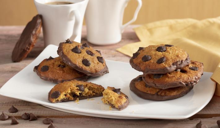 Fotografia em tons de amarelo em uma bancada de madeira com um pano amarelo, um recipiente quadrado branco com alguns cookies de cenoura recheado com chocolate. Ao fundo, duas xícaras brancas com café.
