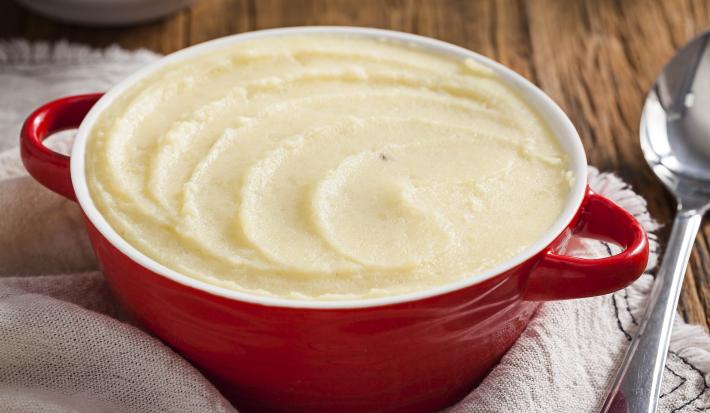 Foto de uma panela vermelha cheia de purê de batatas, acima de um pano bege e ao lado uma colher de servir prateada