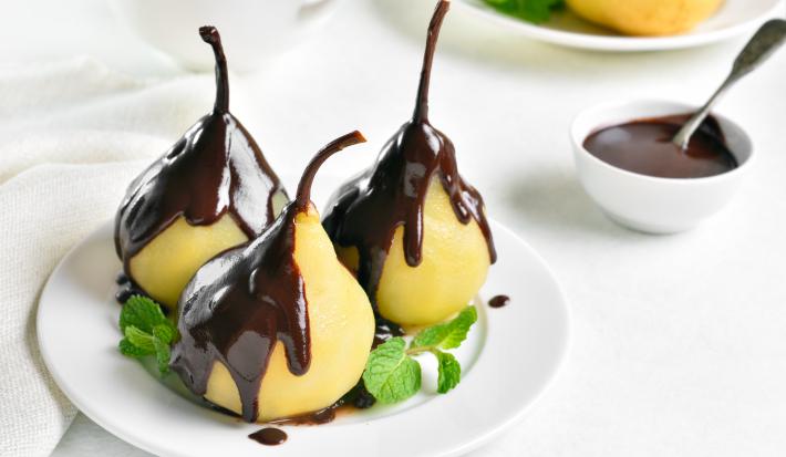 Fotografia de uma sobremesa de peras cozidas no suco de laranja com uma cobertura de chocolate. A sobremesa está dentro de um prato pequeno de vidro com pequenas folhas de hortelã, sobre uma mesa branca. Ao fundo, um pote pequeno com calda de chocolate.
