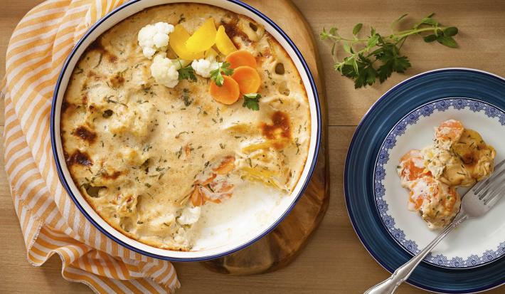 Fotografia em tons de laranja e azul em uma bancada de madeira com um pano com listras laranjas, uma tábua de madeira com um refratário grande e redondo com legumes gratinados. Ao lado, um prato branco com detalhes azuis, com um pedaço dos legumes.
