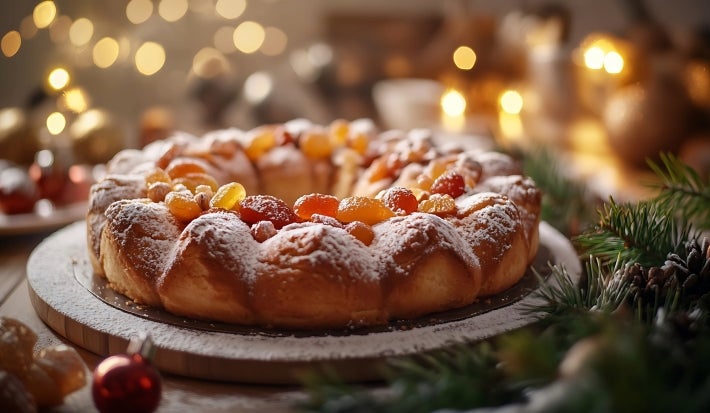 Fotografia de um bolo natalino circular, coberto com açúcar de confeiteiro e decorado com frutas secas coloridas. O fundo apresenta luzes suaves e elementos festivos.