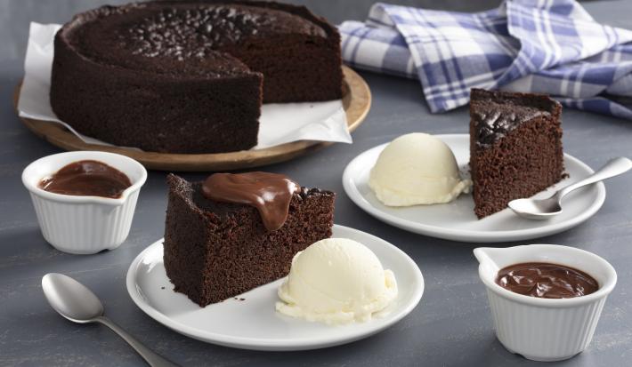 Fotografia em tons de marrom e azul de uma bancada cinza, ao centro dois pratos brancos com fatias de bolo de chocolate e ao lado uma bola de sorvete de creme. Ao fundo o bolo inteiro sem duas fatias.