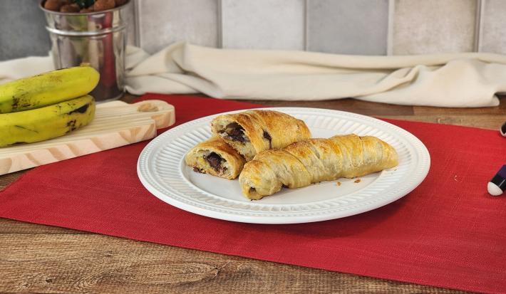 Fotografia em tons de vermelho em uma bancada de madeira escura com um pano vermelho e um prato branco redondo ao centro com duas bananas enroladas em massa folhada e recheadas de chocolate em cima. Ao fundo, cacho de banana e paninho bege.