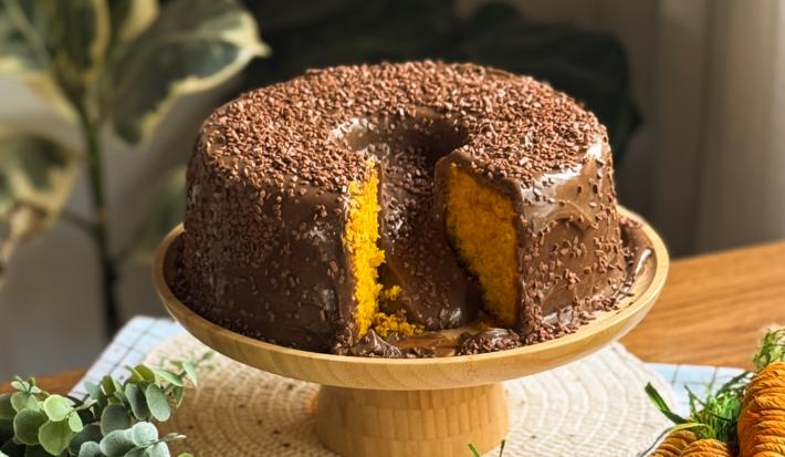 Fotografia de um bolo vulcão de cenoura com brigadeiro. O bolo está com uma fati cortada e está sobre um apoio decorativo de tom amadeirado.