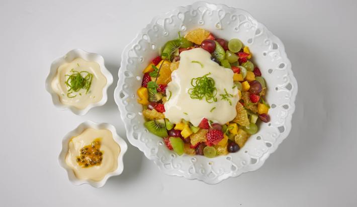 foto em tons de branco de uma bancada branca vista de cima, contém um recipiente redondo branco ao centro com pedaços de frutas e creme. Ao lado dois potinhos redondos brancos com creme.
