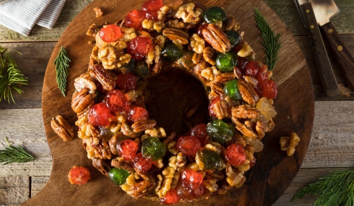 Fotografia de um bolo de frutas circular em forma de guirlanda, adornado com frutas cristalizadas e nozes, colocado sobre uma tábua de madeira rústica, cercado por ramos de pinheiro, evocando um clima natalino e aconchegante.