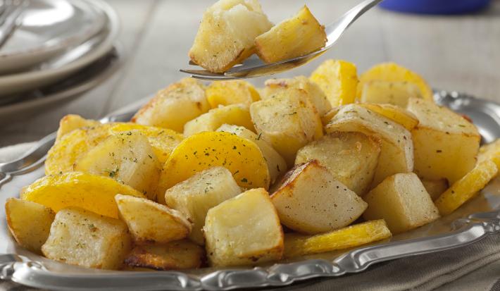 Fotografia em tons de amarelo em uma bancada de madeira com um recipiente de prata com várias mandioquinhas salteadas.