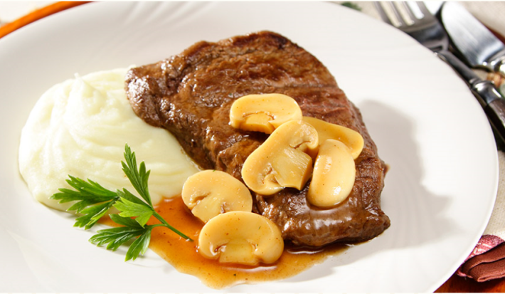 Fotografia em tons de branco e marrom, com bife com molho de champignons acompanhado de purê de batatas e ramo de salsa em prato branco, sobre sous plat marrom, ao lado guardanapo dobrado com garfo e faca por cima, tudo sobre toalha cinza.