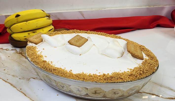 Foto da receita de Torta Banoffee de Travessa, servida em uma travessa oval de vidro, decorada com chantilly batido e biscoitos tostines triturados, tudo em uma bancada de mármore com algumas bananas ao fundo e um tecido vermelho