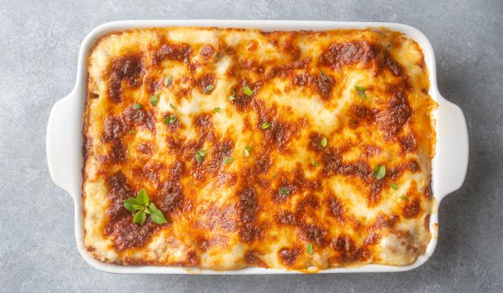 Fotografia vista de cima de uma receita com mandioca, frango e molho de tomate com um gratinado por cima de queijo dentro de uma travessa branca de vidro retangular. O recipiente está sobre uma mesa de cimento queimado.